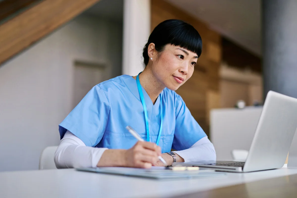 Woman in scrubs making notes and looking at a laptop